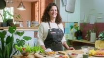 Cheerful mature woman in apron standing by table with fresh food ingredients, posing for camera and smiling in the kitchen. Video portrait, medium shot
