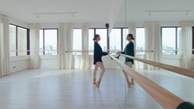 Young girl standing at the barre and doing ankle rotation exercise while warming up feet before ballet class in modern dance studio. Full shot, copy space
