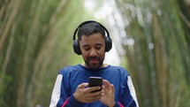 Cheerful young walking on city park with headphones and modern smartphone in hands. Black man reading some good and smiling. Technology for lifestyles.
