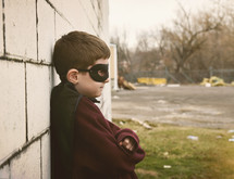 a little boy in a cape and mask leaning against a wall 