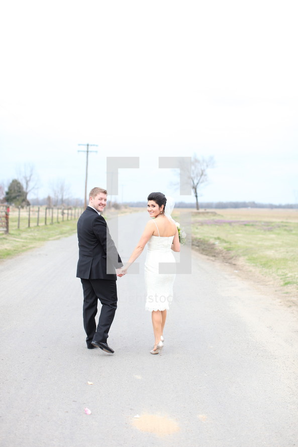 bride and groom holding hands walking down a country road