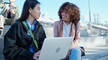 Young African-American and Asian businesswomen using laptop and discussing work, sitting together at outdoor public space in the city. Medium shot, man drinking coffee and using phone in the background
