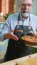 Senior food blogger in apron holding pan with steaming hot stew and commenting recipe on camera, giving online masterclass or filming vlog in kitchen. Vertical clip
