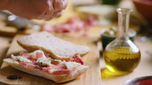 Hands of woman adding fresh basil leaves to gourmet sandwich with prosciutto and blue cheese on ciabatta bread. Close-up view, selective focus
