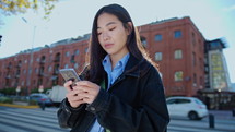 Young beautiful Asian woman standing on the street in the city, text messaging on smartphone and smiling. Medium handheld shot, lens flare
