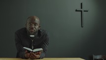Black Priest Holding Opened Bible and Saying Prayer in Church