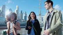 Company of young multi-ethnic coworkers walking together on the riverbank in downtown, smiling and having discussion, spending time outdoors on sunny day. Medium shot, cityscape in the background
