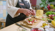 Mature woman in apron adding fresh basil leaves to gourmet sandwich, then enjoying the smell and commenting on camera, filming cooking vlog or video recipe. Handheld tilt-up shot
