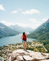 woman standing on a cliff overlooking a town by a mountain lake 
