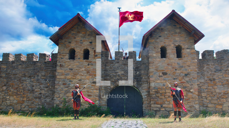 Roman soldiers guarding a military fort on a cloudy summer day. The flag of the Roman Empire fluttering on the crenellated wall between the watchtowers.