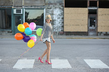 Woman in a hat and high heels walking down the street holding balloons.