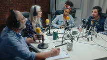 Podcast co-hosts in headphones sitting around the table with mics and web cameras on tripods, discussing topic at recording studio with professional audio equipment and TV screen