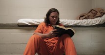 A sad man, convicted prisoner inmate wearing an orange prison uniform/jumpsuit sitting in his prison cell reading a bible.