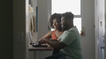 African couple spend time in cozy kitchen, wife sit at bar counter use laptop discuss with husband retail purchase, browse internet having fun online. Modern wireless tech, order, buy remotely concept
