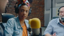 Senior man in headphones sitting at desk at studio, speaking into mic and making notes on paper when recording audio podcast