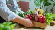 Male chef bringing wooden crate full of assorted fresh vegetables and placing eggplant, artichoke and bell peppers on kitchen table before cooking. Close-up shot
