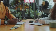 Podcast Co-Hosts Having Discussion at Desk with Mics and Planners in Studio
