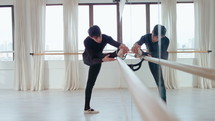 Full shot of young male ballet dancer stretching with one leg on the barre in dance studio with mirror wall
