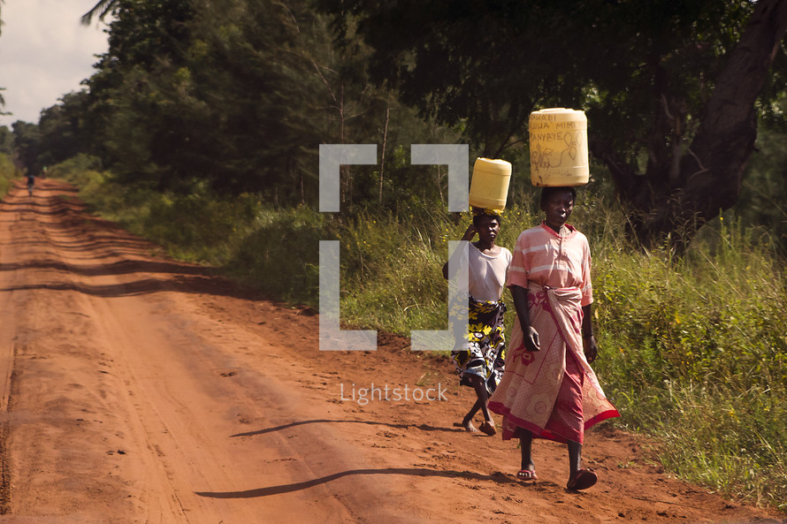 ladies carrying water on their heads walking on a dirt road 