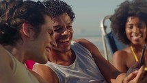 Happy smiling adults friends laughing outside at the beach at something in smartphone or mobile phone. Young multiracial people spending time together. Friendship, communication, youth and lifestyle concept