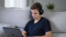 Teenager using notebook studying at the home living room and sits on couch. Young guy works on notebook, sends messages, makes online purchases, watching movies, working.
