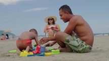 Family together at the beach. Father plays on the sand with his son while mom takes a sunbath on a family summer vacation
