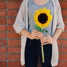 A woman stands against a brick wall holding a large sunflower.