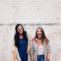 Two laughing young women against a white brick wall.