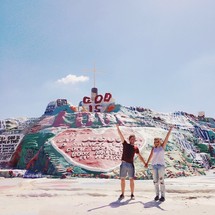 Couple holding hands with arms raised standing in front of a Salvation Mountain in Slab City, CA.