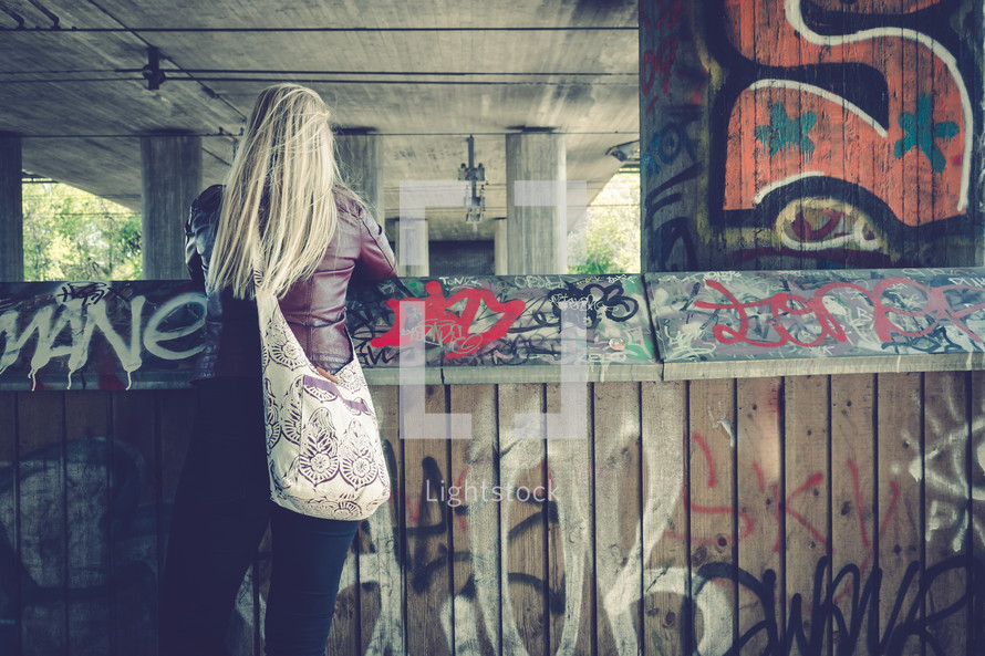 young woman looking over a graffiti covered wall under an overpass 