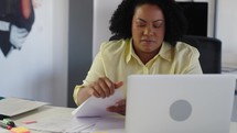Young busy black business woman working on project paperwork at laptop.