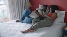 Happy young couple in bed on their phone on social media, browsing internet and online meme. Black couple in bedroom with smartphone. Love, relationship and relax 