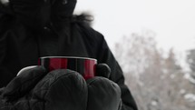Person outside wearing a black coat and gloves with a mug of steaming beverage