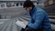 a man reading a Bible on steps in a city 