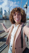 Young cheerful African-American female blogger standing outdoors on the riverside, showing the city and chatting on camera, filming vlog or web calling. Vertical POV shot
