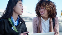 Young African-American and Asian female colleagues smiling, discussing business, using laptop, spending day outdoors at public space in the city. Medium close-up shot
