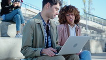 Young man sitting outdoors with laptop, talking with African-American female colleague at urban public space in the city. Medium shot
