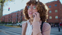 Young African American businesswoman speaking on mobile phone standing on city street on sunny day. Medium shot, lens flare
