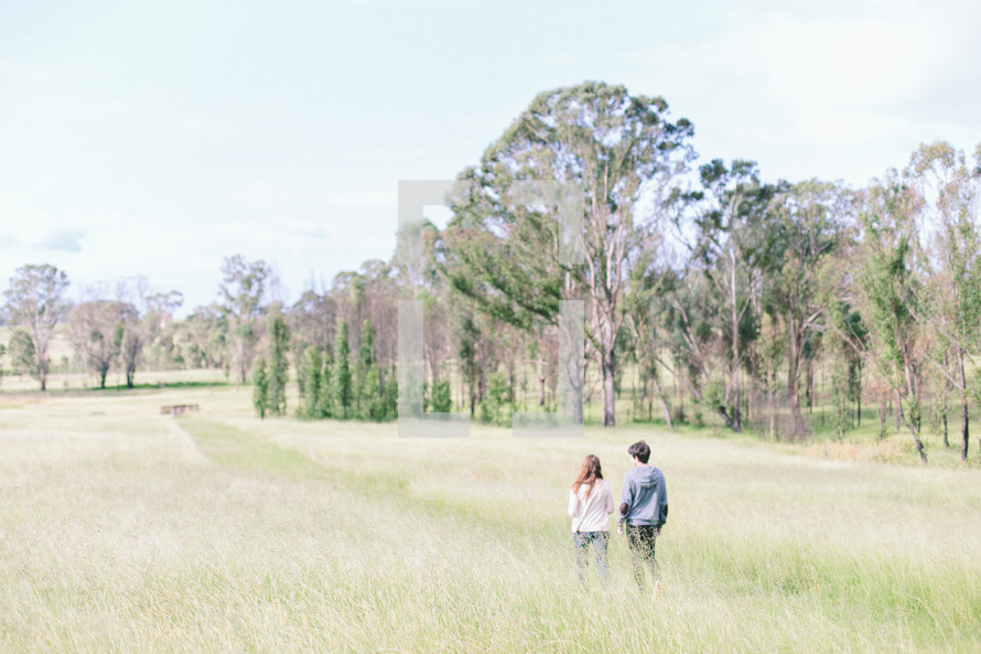 Couple walking in a grassy field near a tree line.