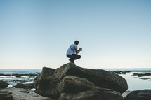man squatting on a rock on shore holding a camera 
