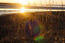 A woman sits in tall grass facing the sunset.