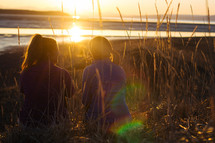 Two girls sit in tall grass facing a glowing sunset.