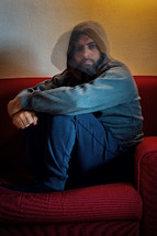 Depressed man, with hood on his head, sitting alone on a sofa