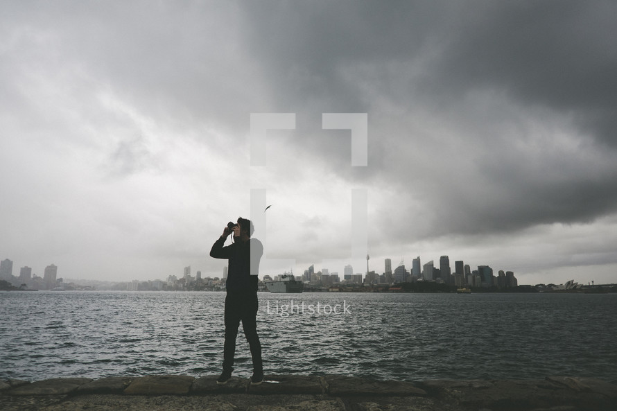 man standing across the river from a city taking a picture 