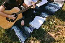 young women sitting on a blanket outdoors reading Bibles and playing a guitar 