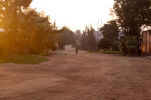 Boy walking down a dirt road in a town, carrying a bag.