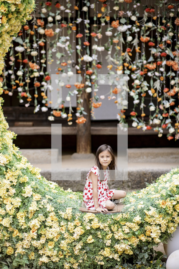 A young girl sitting in a crescent moon with a flower wall behind her