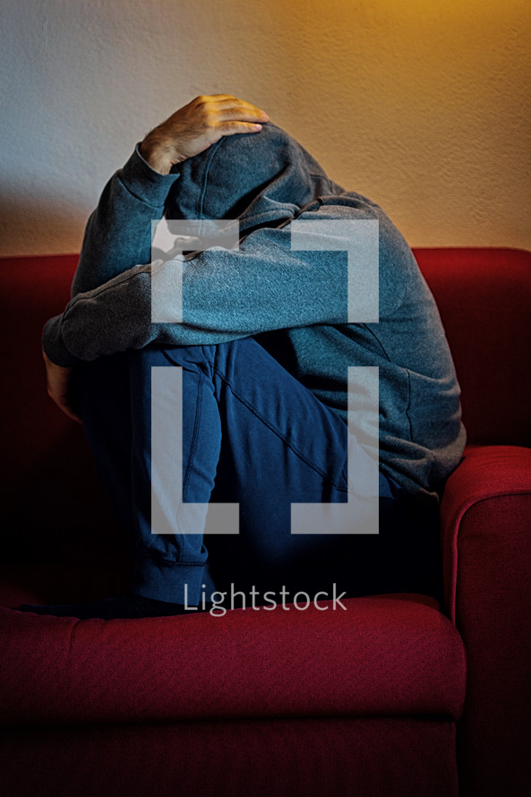 Young man is sitting alone on a couch, curled up with his head in his hands, suffering from depression