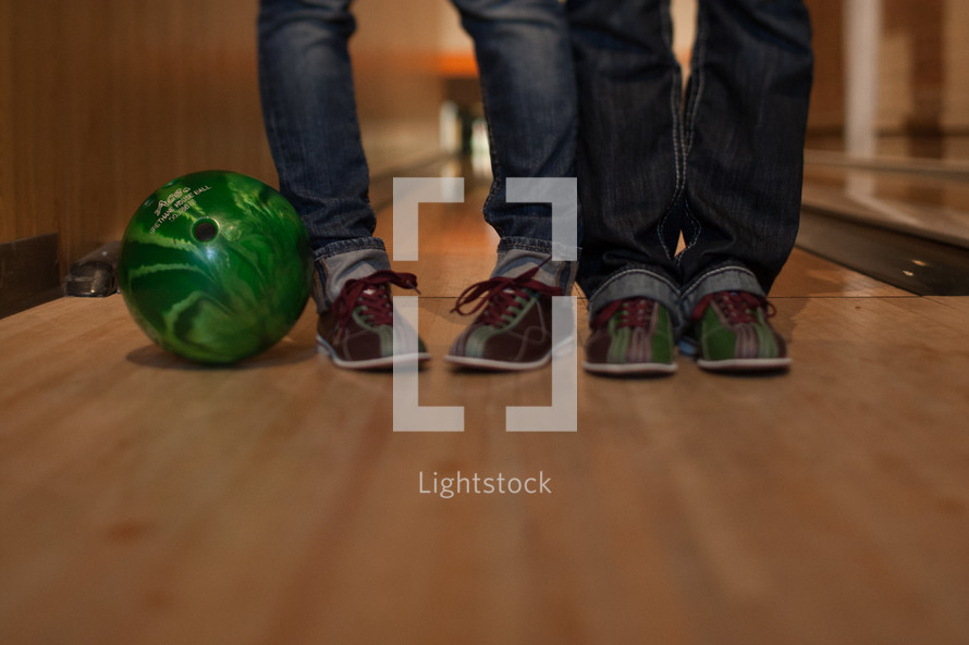 Two pairs of legs in jeans  and bowling shoes standing on wood floor with green bowling ball.