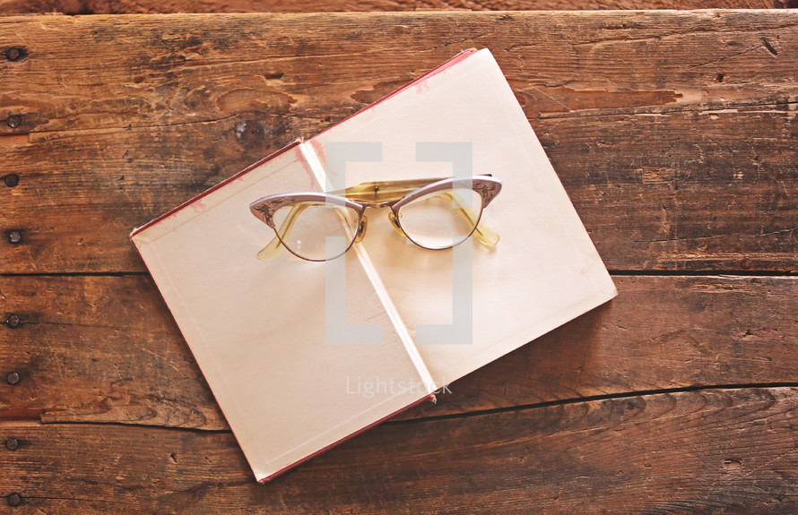 Eye glasses on an open book, which is on a rugged wooden table.
